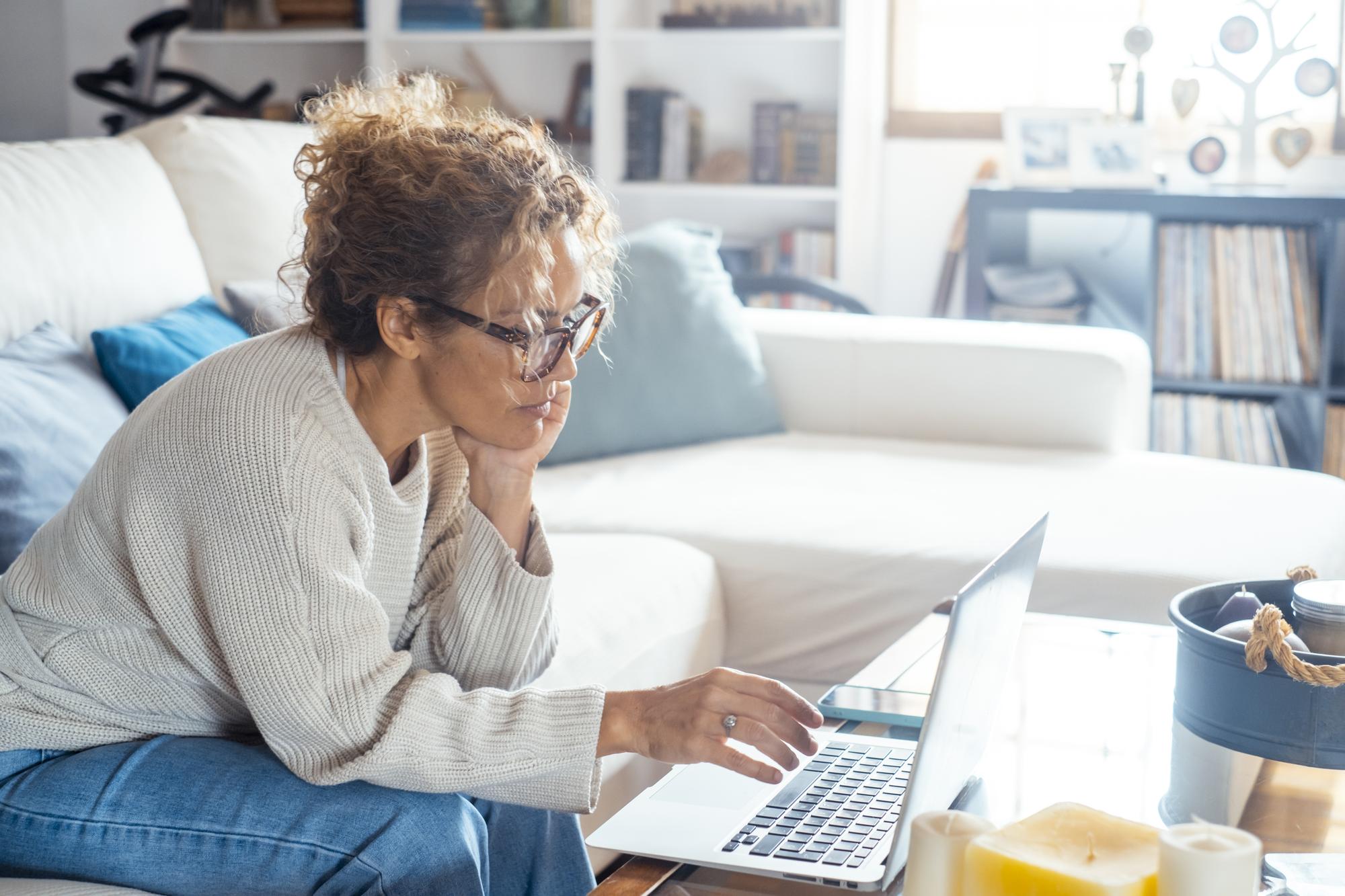 Vue latérale d'une femme seule à la maison navigant sur les pages Web d'Internet en utilisant un ordinateur portable assis sur le canapé Les femmes modernes travaillant sur l'ordinateur dans le travail à distance lieu de travail de bureau alternatif Site Web d'adhésion